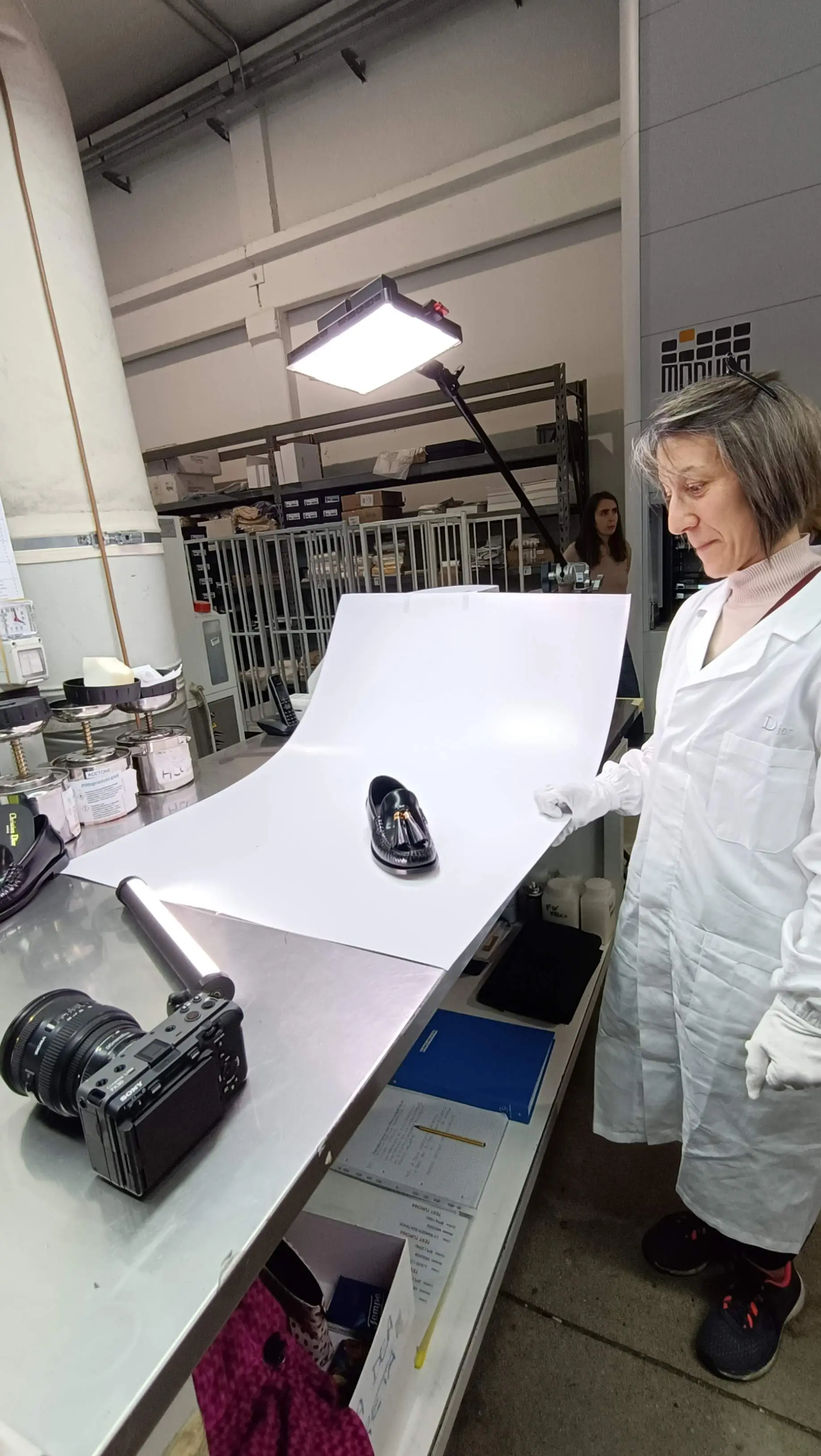 Artisan in a white coat at a lit table during a Dior savoir-faire shoot, with the camera rigged low beside the bench