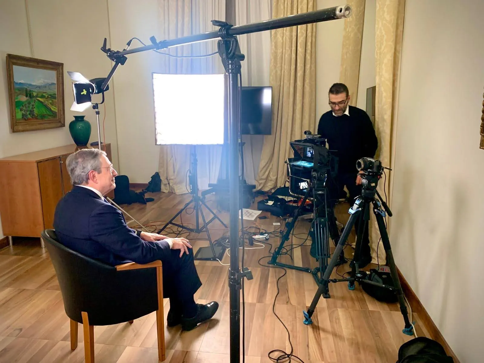 Wide view of the improvised interview set at the Bank of Italy: Mario Draghi in an armchair against a bare wall, two cameras on tripods, boom microphone, LED panel and the prompter carrying the remote interviewer