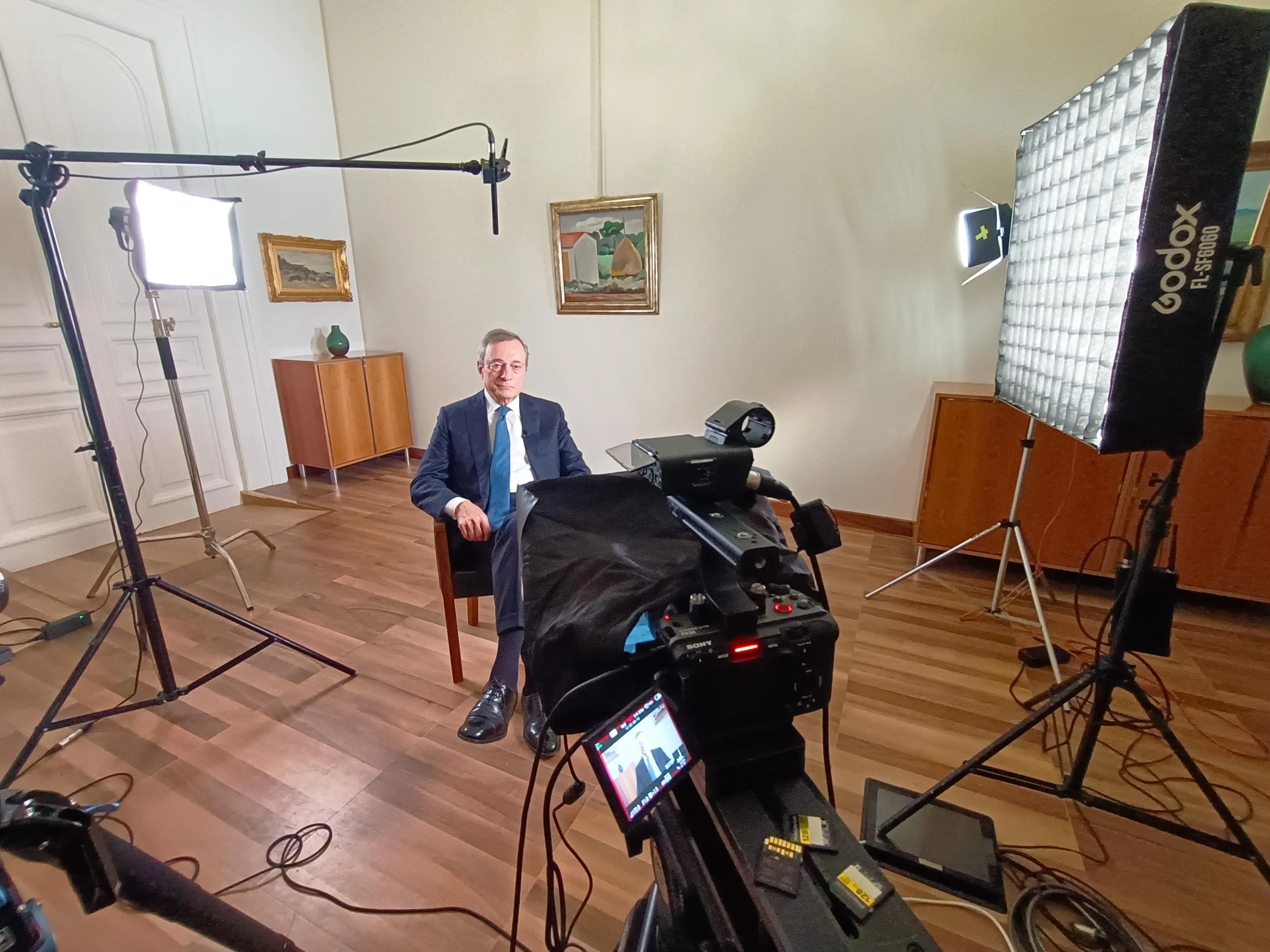 Mario Draghi seated for the interview in a plain room at the Bank of Italy, framed from behind the Sony FX30, with a Godox FL-SF6060 softbox on the right, a second LED panel on the left and a boom microphone overhead