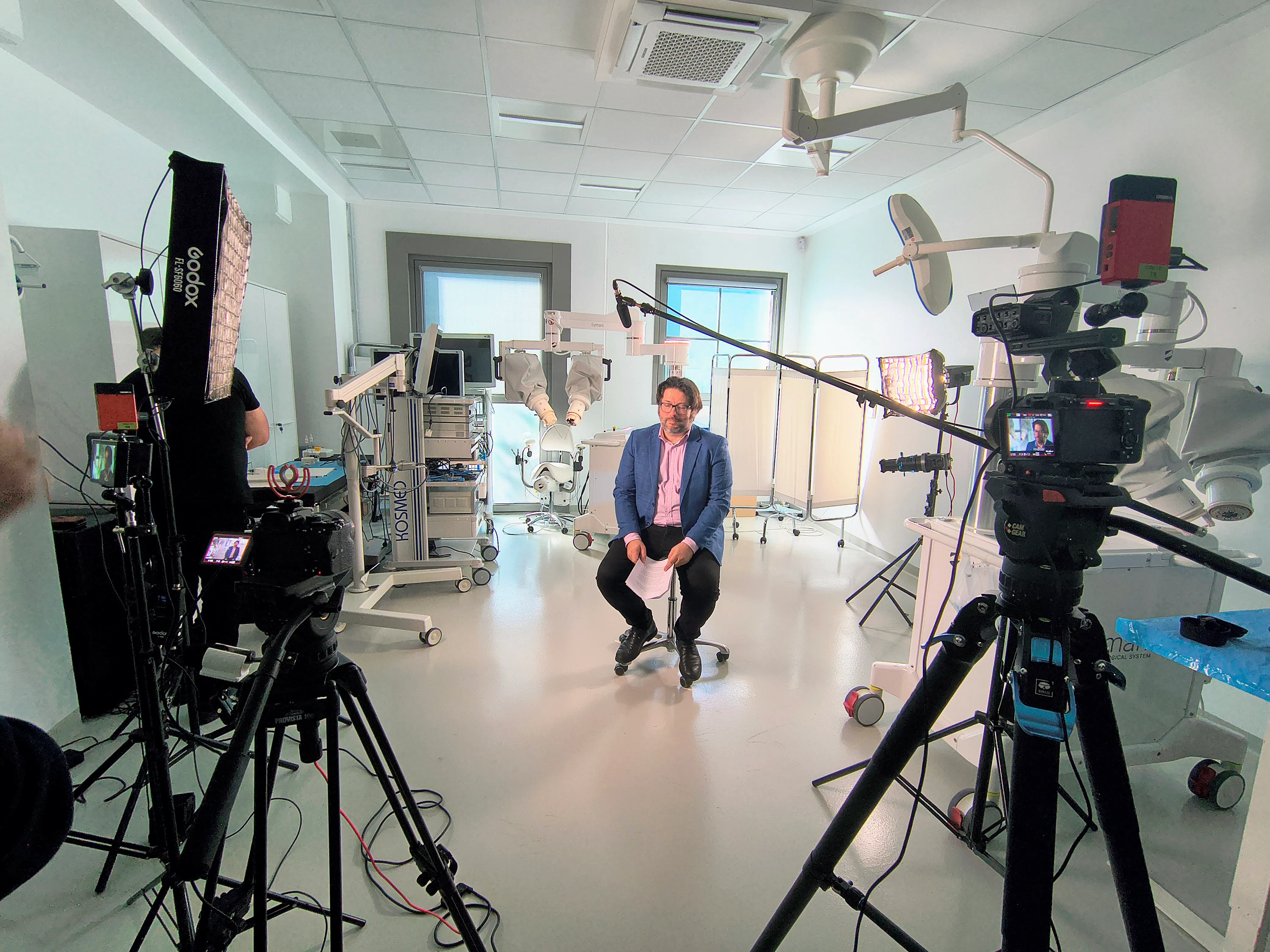 Two-camera interview setup inside an operating theatre with robotic surgery equipment, lit with softbox and LED panels