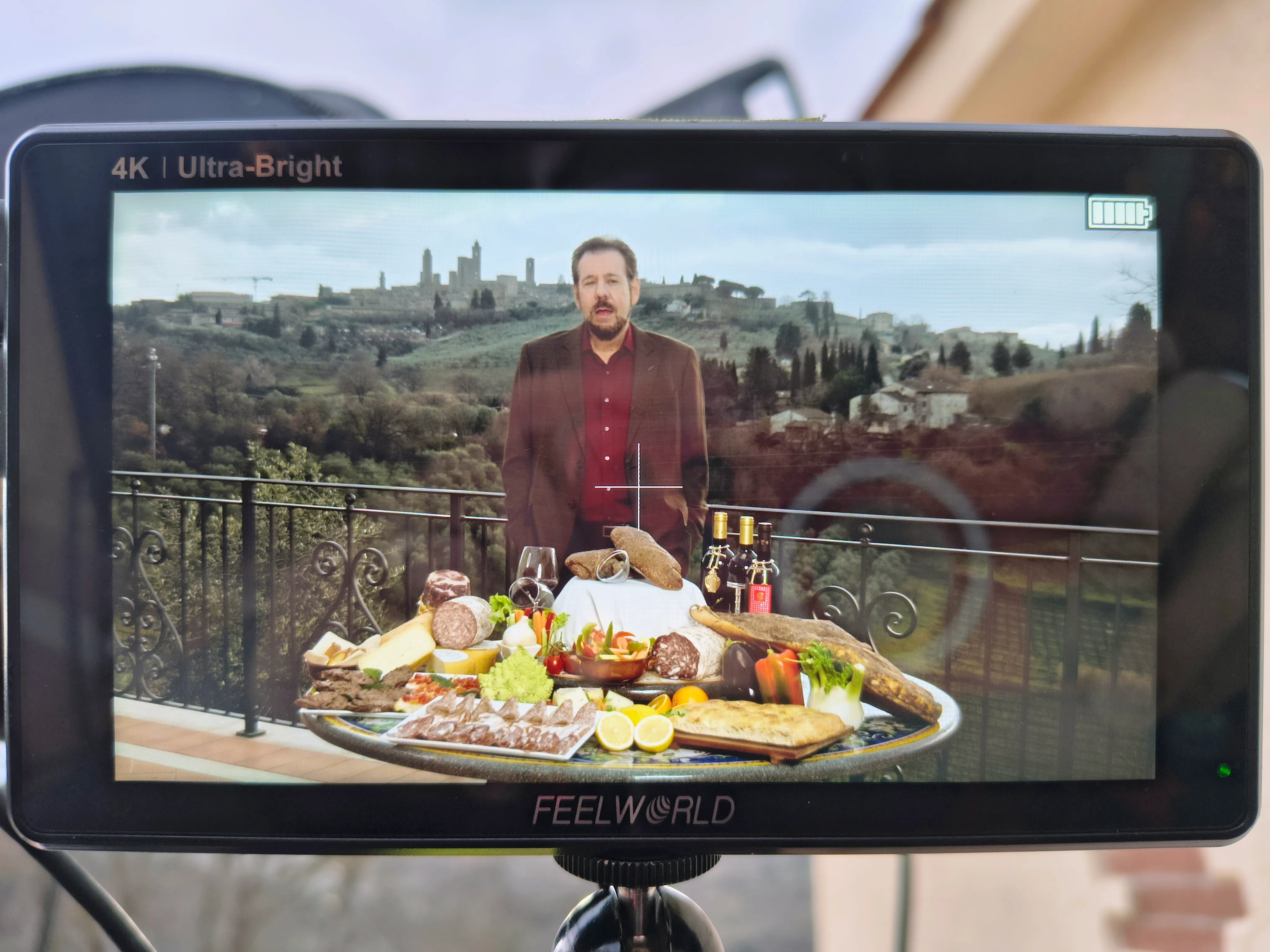 The field monitor during the live stream, with the presenter framed behind the food and the towers of San Gimignano in the background