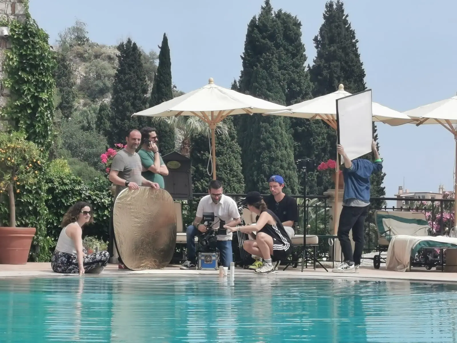 Film crew on a Dior shoot beside the pool of a hilltop terrace in Taormina, Sicily, with a gold reflector and a white bounce board set up among parasols and cypresses